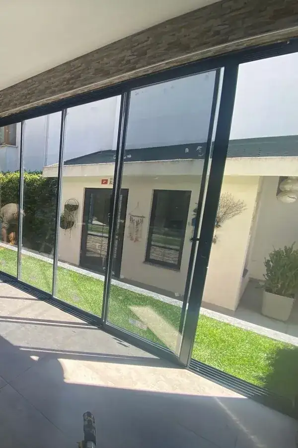 Bahçeye açılan tavana kadar sürme cam duvar; zemindeki ray ve siyah ince profiller