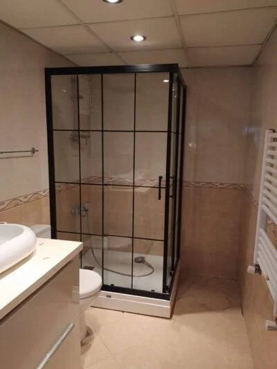 Banyoda siyah çerçeveli, ızgara desenli köşe duşakabin; önde lavabo, sağda havlupan